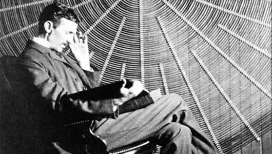 An image of Nikola Tesla sitting and reading in front of a spiral coil of his high-voltage transformer.